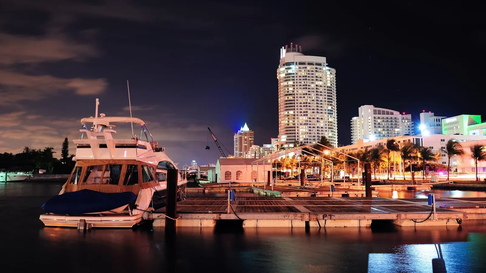 Yacht moored at a marina at night, with illuminated high-rise buildings behind.