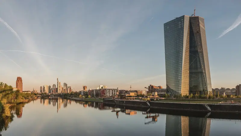 The glass tower of the European Central Bank beside the river Main in Frankfurt at dawn, with the city skyline and its reflection in the water.