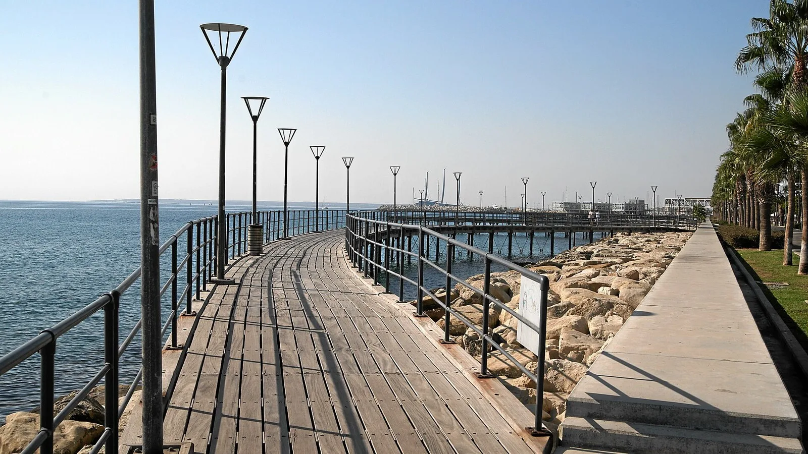 Promenada de lemn de pe faleza din Limassol, Cipru, curbată de-a lungul mării într-o zi senină, cu stâlpi de iluminat, stânci și palmieri alături.